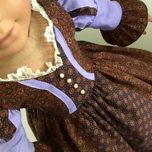 Puede incluir: Una mu&ntilde;eca con un vestido marr&oacute;n con estampado floral con detalles y mangas color lavanda. El vestido tiene un cuello de encaje blanco y botones perlados. La piel de la mu&ntilde;eca es de color melocot&oacute;n p&aacute;lido.