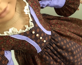 May include: A doll wearing a brown floral print dress with lavender accents and sleeves. The dress features a white lace collar and pearl-like buttons. The doll's skin is a pale peach color.