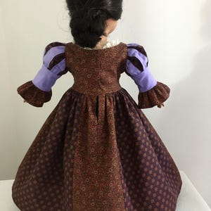 Puede incluir: Mu&ntilde;eca con un vestido marr&oacute;n y morado estampado. El vestido tiene una falda amplia y mangas abullonadas en morado y marr&oacute;n. La mu&ntilde;eca tiene el pelo oscuro peinado con una trenza. El vestido recuerda a la vestimenta hist&oacute;rica.