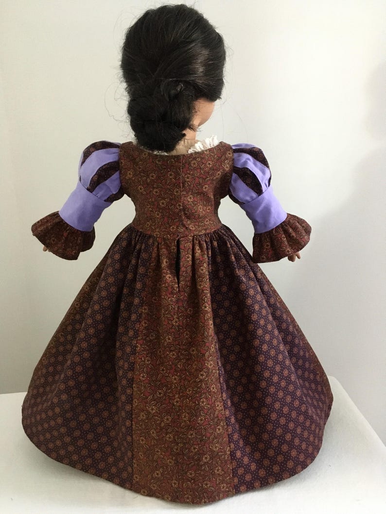 Puede incluir: Mu&ntilde;eca con un vestido marr&oacute;n y morado estampado. El vestido tiene una falda amplia y mangas abullonadas en morado y marr&oacute;n. La mu&ntilde;eca tiene el pelo oscuro peinado con una trenza. El vestido recuerda a la vestimenta hist&oacute;rica.
