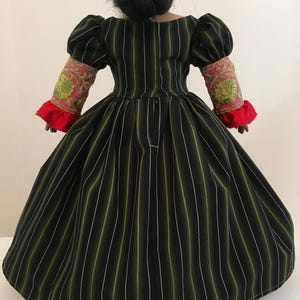 May include: A doll wearing a black and green striped dress with white and olive green stripes. The dress has puffed sleeves with pink and gold embroidered cuffs and red ruffled trim. The doll's hair is styled in a bun.