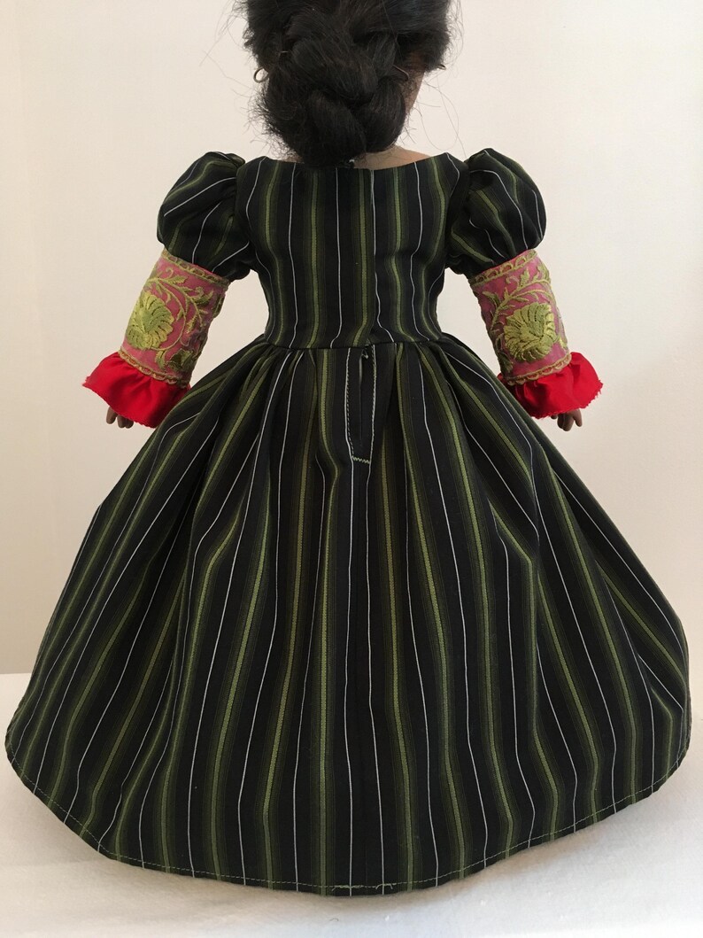 May include: A doll wearing a black and green striped dress with white and olive green stripes. The dress has puffed sleeves with pink and gold embroidered cuffs and red ruffled trim. The doll's hair is styled in a bun.