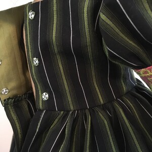 May include: Close-up of a black dress with vertical green and white stripes. The dress features a high collar with silver buttons and a green lining. The sleeve cuff is pink with green embroidery.