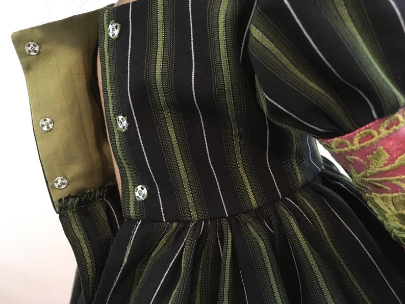 May include: Close-up of a black dress with vertical green and white stripes. The dress features a high collar with silver buttons and a green lining. The sleeve cuff is pink with green embroidery.