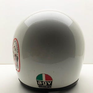 May include: A white motorcycle helmet with a glossy finish. The helmet has a black trim and features two stickers: one with a red, white, and black design and text, and another with the Italian flag and the letters "agv".