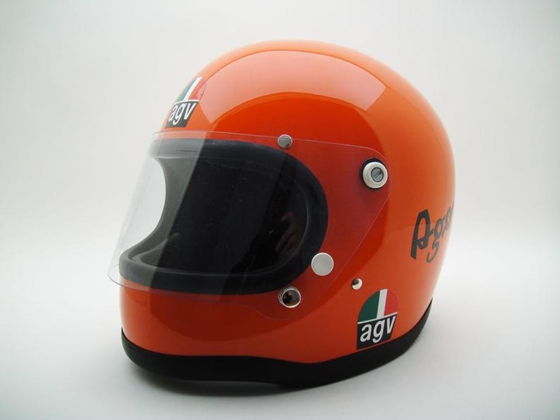May include: Glossy orange motorcycle helmet with a clear visor and black interior. Features the "AGV" logo and script on the side. The helmet has a black trim around the base and a clear visor.