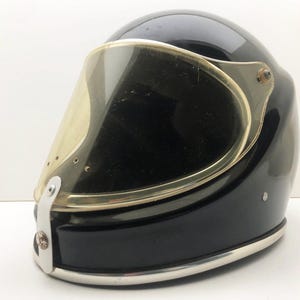 Può includere: Casco da moto nero lucido con visiera trasparente e curva. Il casco ha un bordo argentato sul bordo inferiore e intorno alla visiera. La visiera è fissata con hardware argentato. Il casco è progettato per la sicurezza e la protezione.