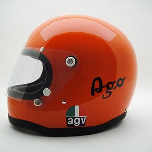 May include: Orange motorcycle helmet with a clear visor. The helmet has the word "Ago" in black script on the side, and the "AGV" logo with Italian flag colors. The helmet is a classic design.