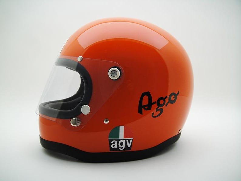 May include: Orange motorcycle helmet with a clear visor. The helmet has the word "Ago" in black script on the side, and the "AGV" logo with Italian flag colors. The helmet is a classic design.