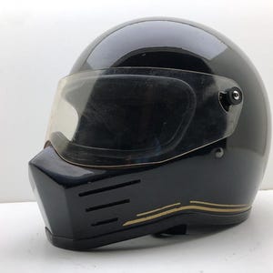 May include: A glossy black full-face motorcycle helmet featuring a clear visor, a prominent chin bar with three horizontal ventilation slits, and thin gold pinstripe detailing along the lower edge of the shell, presented against a plain background.