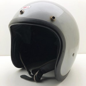 Può includere: Un casco da moto aperto di colore grigio chiaro con un bordo nero lungo il profilo. Il casco presenta un logo Bell rosso e bianco sulla parte anteriore, due bottoni automatici metallici per una visiera e un sottogola nero con chiusura a doppio anello.