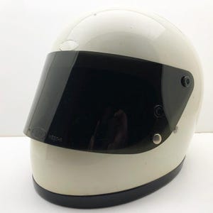 Könnte beinhalten: Cremefarbener Motorradhelm mit glänzendem Finish und schwarzem Visier. Der Helm hat eine runde Form und einen dunklen Rand an der Basis. Das Visier ist mit silbernen Nieten befestigt.