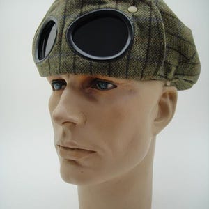 Può includere: Un cappello di lana a quadri verde e marrone con due occhiali neri sul davanti. Il cappello ha un piccolo bottone bianco sulla parte superiore.