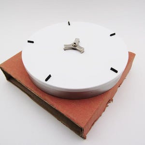 Könnte beinhalten: Ein weißes, kreisförmiges Objekt mit schwarzen Strichen und einem silbernen Metallteil obenauf, das auf einer rotbraunen Kartonbasis ruht. Das Objekt scheint ein Zifferblatt oder eine ähnliche Komponente zu sein.