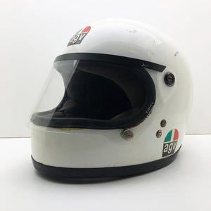 May include: White motorcycle helmet with a clear visor and black trim. The helmet features the "agv" logo in red, white, and green. This full-face helmet is designed for safety and protection, a classic design.