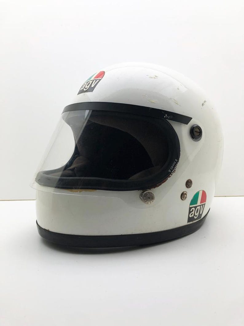 May include: White motorcycle helmet with a clear visor and black trim. The helmet features the "agv" logo in red, white, and green. This full-face helmet is designed for safety and protection, a classic design.
