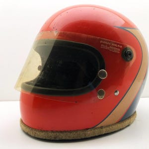 Könnte beinhalten: Ein glänzender roter Integralhelm mit einem beigefarbenen und dunkelblauen Seitenstreifen. Der Helm verfügt über ein getöntes Visier mit dem Aufdruck STAR FLITE MODEL FC-1B an der Seite. Er hat einen beigen Stoffrand am unteren Rand.