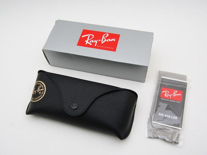 Puede incluir: Un estuche de gafas de cuero negro con un logotipo dorado, una caja gris con un logotipo rojo y un pa&ntilde;o de limpieza con un logotipo rojo. El estuche se cierra con un broche. La caja y el pa&ntilde;o tienen el logotipo de Ray-Ban.
