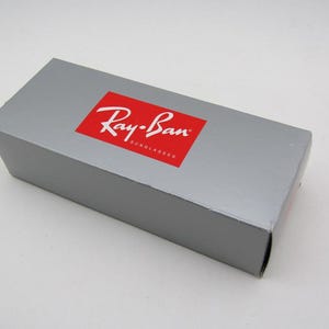 Puede incluir: Una caja rectangular gris con un logotipo rojo de Ray-Ban. La caja es para gafas de sol y tiene el nombre de la marca en blanco. La caja est&aacute; cerrada y se encuentra sobre una superficie blanca.