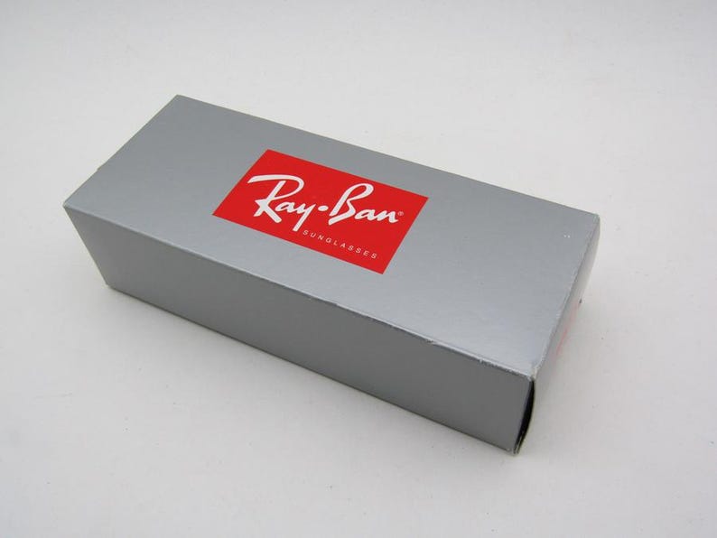 Puede incluir: Una caja rectangular gris con un logotipo rojo de Ray-Ban. La caja es para gafas de sol y tiene el nombre de la marca en blanco. La caja est&aacute; cerrada y se encuentra sobre una superficie blanca.