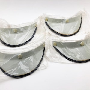 以下が含まれることがあります： 黒い縁取りと小さなボタンが付いた、グレーのバイザーが4つ。それぞれ透明なプラスチックで個別に包装されています。バイザーは湾曲したデザインで、おそらく日焼け防止用です。背景は白です。