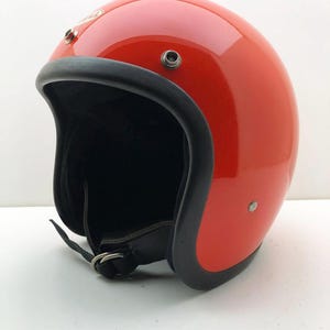 Può includere: Un casco da moto aperto di colore rosso brillante lucido con un bordo in gomma nera, una fodera interna nera e un cinturino sottogola nero con chiusure a doppio anello in metallo. Il logo del marchio Bell è visibile sulla parte anteriore.