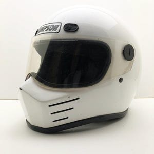 Può includere: Casco da moto bianco con visiera trasparente. Il casco ha la scritta "Simpson" stampata sul davanti. Il casco ha un bordo nero e tre fessure di ventilazione orizzontali sul mento.