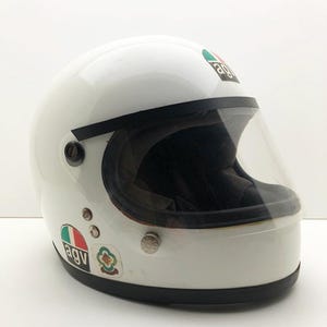 May include: A white vintage motorcycle helmet with a clear visor and black edging. The helmet features several stickers, including the brand logo. A classic design for motorcycle enthusiasts.