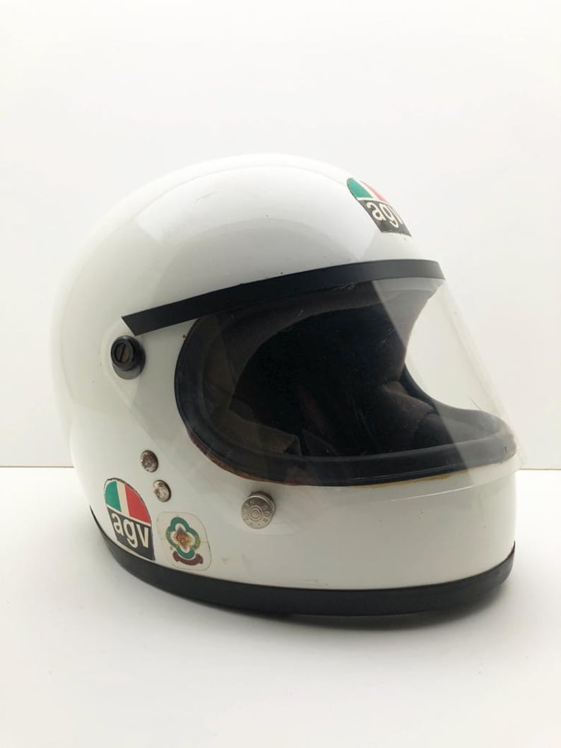 May include: A white vintage motorcycle helmet with a clear visor and black edging. The helmet features several stickers, including the brand logo. A classic design for motorcycle enthusiasts.