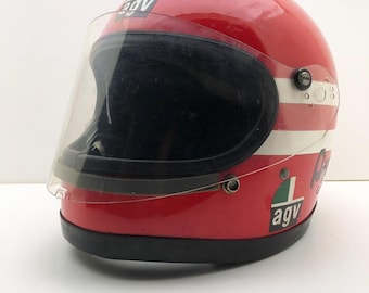 May include: A vintage red motorcycle helmet with a clear visor. The helmet features white stripes and the "agv" logo in green and white. The helmet has a black trim and a clear visor.