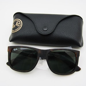 Puede incluir: Gafas de sol marrones oscuras con un estuche negro. Las gafas tienen una montura cuadrada y lentes oscuras. El estuche es negro, texturizado y tiene un cierre de bot&oacute;n. El logotipo de Ray-Ban es visible en las gafas y el estuche.