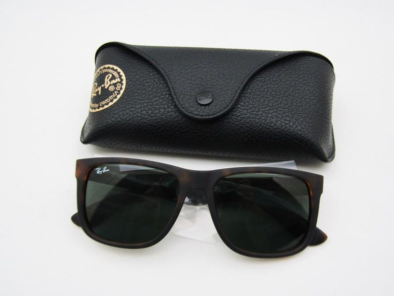 Puede incluir: Gafas de sol marrones oscuras con un estuche negro. Las gafas tienen una montura cuadrada y lentes oscuras. El estuche es negro, texturizado y tiene un cierre de bot&oacute;n. El logotipo de Ray-Ban es visible en las gafas y el estuche.