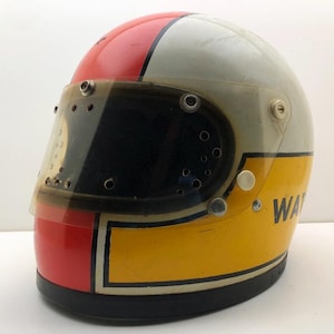 Può includere: Un casco da moto integrale con base bianca, una prominente striscia centrale rossa e una sezione inferiore gialla bordata di nero. Il casco include una visiera trasparente oscurata con diversi fori di ventilazione e il testo parziale WAT visibile.