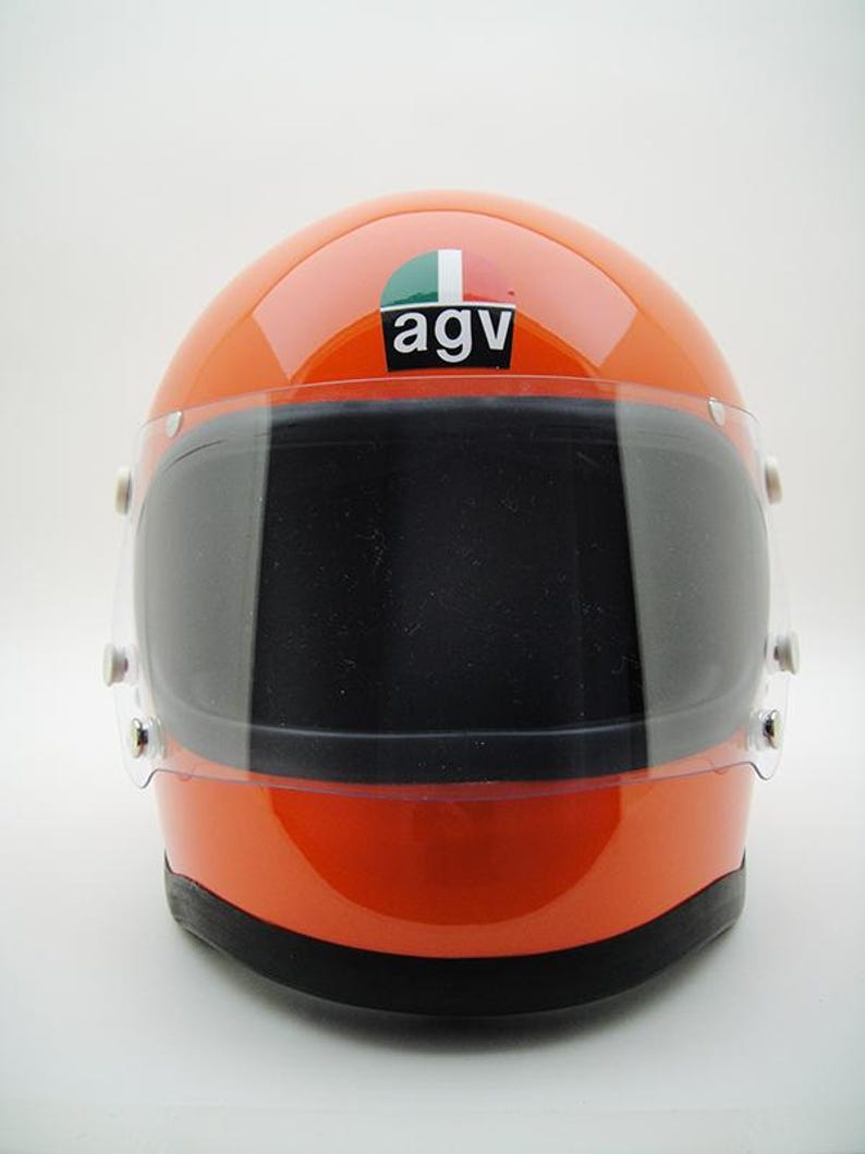 May include: Orange motorcycle helmet with a dark visor. The helmet has the logo "agv" on the top. The visor is clear and has a black tint. The helmet is a glossy orange color.
