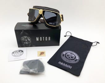 May include: Motorcycle goggles with a gold frame and dark lenses, resting on a black box with the word "MOTOR". Includes a black drawstring bag and accessories. The goggles have a brown leather strap.