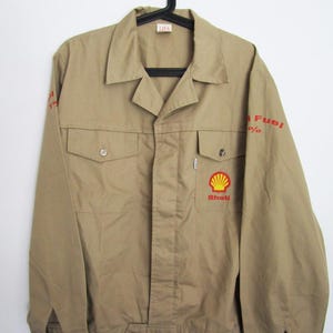 May include: Tan work jacket with a button-up front, two chest pockets, and long sleeves. The Shell logo is on the right chest, and red text on the sleeves reads "Fuel %." The jacket is hanging on a black hanger.
