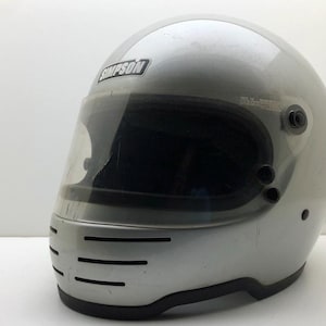 May include: A silver full-face helmet featuring a clear visor and a chin bar with three horizontal ventilation slots. The brand name SIMPSON is printed in black and white on the upper front section of the helmet shell above the visor.