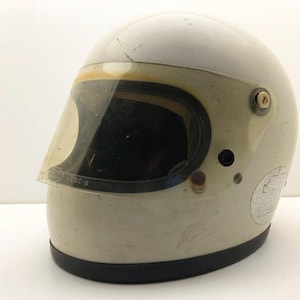 Può includere: Un casco da moto integrale bianco vintage con una visiera trasparente leggermente ingiallita. La visiera riporta la scritta BELL VESC-8. La calotta del casco mostra segni di usura, tra cui piccoli graffi e un adesivo circolare sul lato.