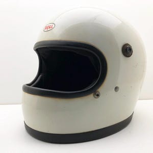 Può includere: Un casco da moto integrale vintage color bianco sporco con bordo nero attorno all'apertura oculare e alla base. Il casco presenta un logo Bell rosso e bianco sulla fronte, una fodera interna scura e componenti metallici laterali per la visiera.