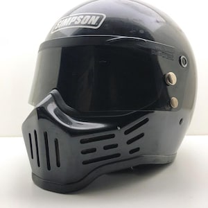 Könnte beinhalten: Schwarzer Motorradhelm mit getöntem Visier und markantem Frontgrill-Design. Der Helm trägt das "SIMPSON"-Logo in Weiß auf der Oberseite. Für Motorradfahren konzipiert.