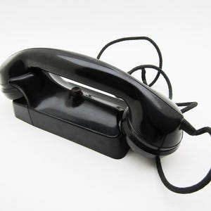 Op de afbeelding: Een vintage zwarte telefoonhoorn met een houder en een opgerold snoer. De glanzende retro-telefoon heeft een gebogen handvat en een kleine knop. Het snoer is zwart en in lussen gelegd. De telefoon staat tegen een witte achtergrond.
