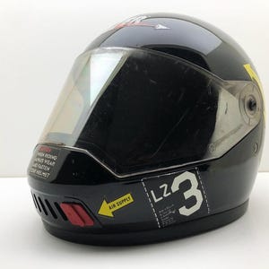 Può includere: Un casco integrale da moto nero lucido con visiera trasparente, una presa d'aria rossa sul mento e adesivi a forma di freccia gialla con la scritta AIR SUPPLY. Sul lato è presente la scritta bianca LZ 3 e un adesivo di avvertenza.
