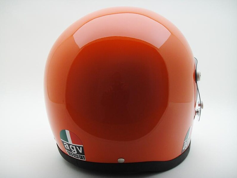 May include: Glossy orange motorcycle helmet with a black trim. The helmet features a reflective surface and a small sticker with the letters "agv" and the colors green, white, and red. The helmet is designed for safety and protection.