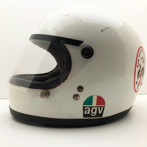 May include: A white vintage motorcycle helmet with a clear visor and black trim. The helmet features a sticker with "BOB HEAD" text, an Italian flag sticker, and the "agv" logo. A classic design.