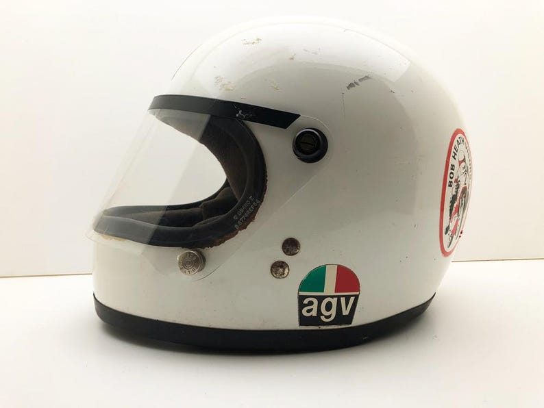 May include: A white vintage motorcycle helmet with a clear visor and black trim. The helmet features a sticker with "BOB HEAD" text, an Italian flag sticker, and the "agv" logo. A classic design.
