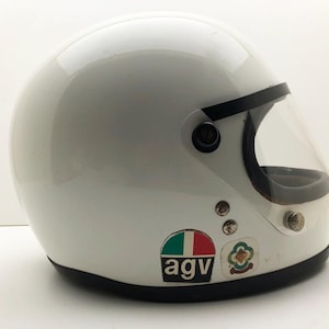 May include: White vintage motorcycle helmet with a clear visor and black trim. The helmet has a glossy finish and features the "agv" logo and other decals. A classic design for motorcycle enthusiasts.