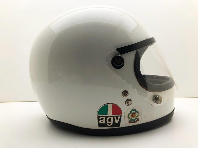 May include: White vintage motorcycle helmet with a clear visor and black trim. The helmet has a glossy finish and features the "agv" logo and other decals. A classic design for motorcycle enthusiasts.