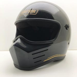 May include: A black motorcycle helmet with a distinctive square chin and a gold "SIMPSON" logo. The helmet has a wide visor opening and a gold stripe. The front features three horizontal vents. The helmet is designed for safety.