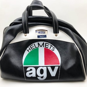 May include: Black and white duffel bag with a white stripe and handles. The bag features the text "HELMETS" above a green and red graphic, and "agv" below. A small tag reads "WHITE".