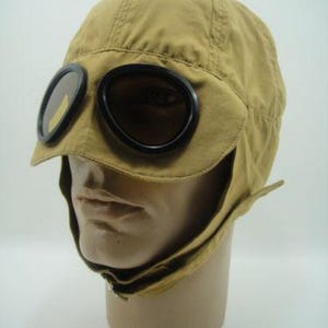 Può includere: Un cappello da aviatore in cotone color beige con visiera nera e occhiali. Il cappello ha una patta che può essere tirata giù per coprire le orecchie e il collo.